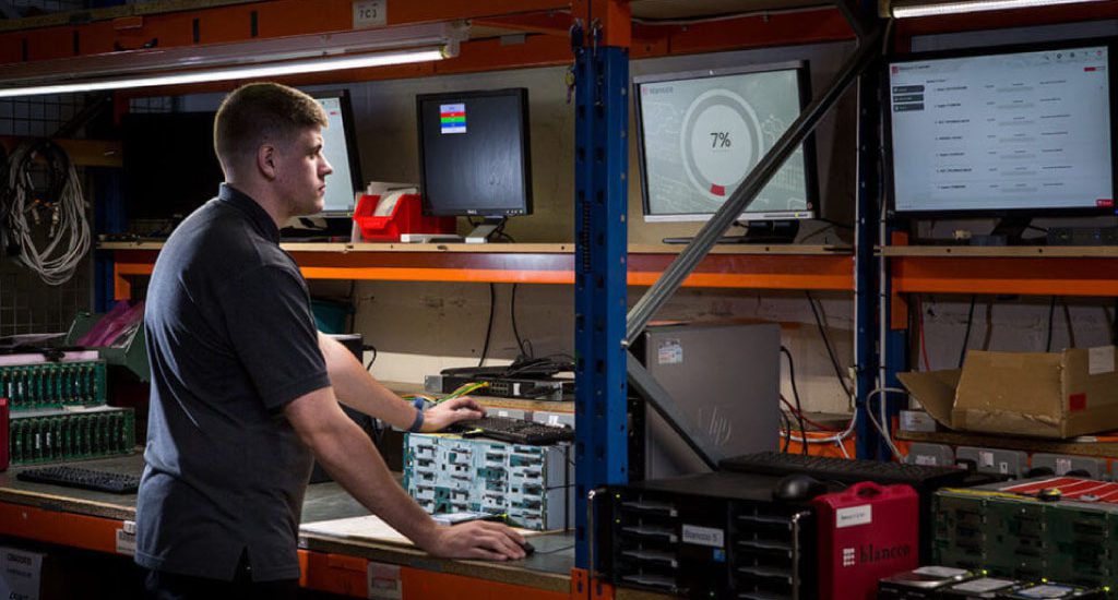 A man wearing a dark polo shirt stands in front of several screens.