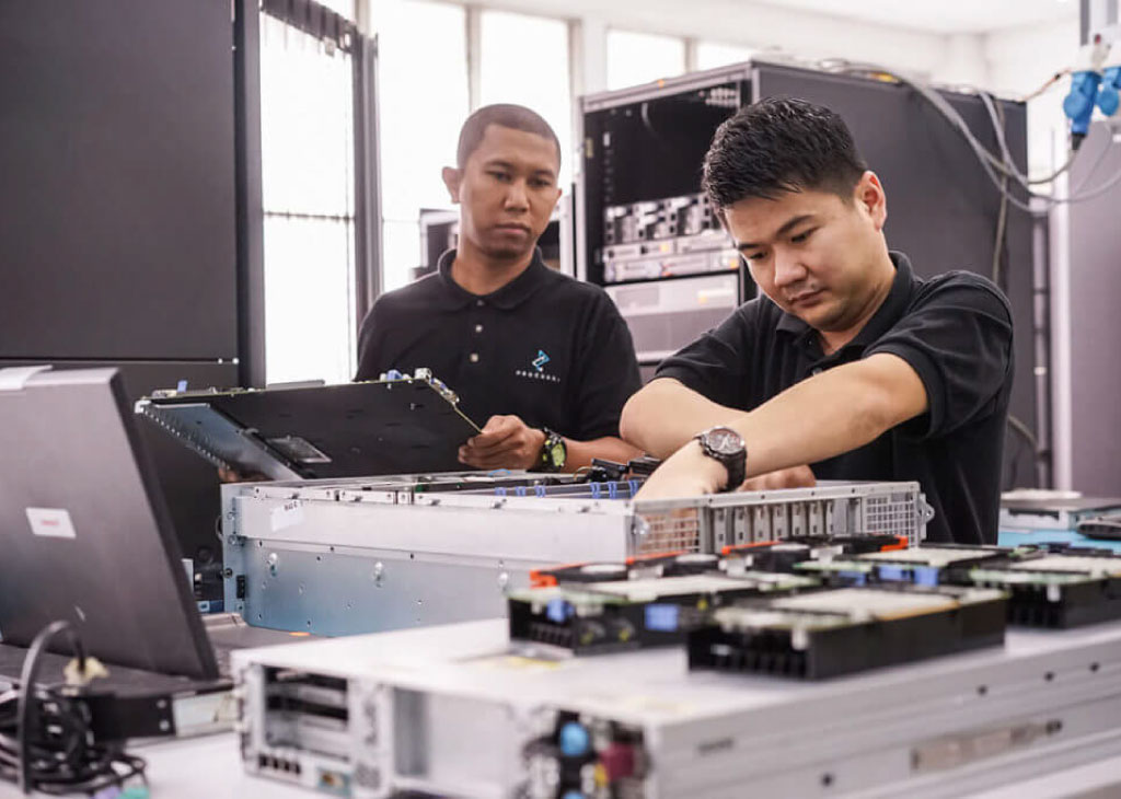 Two male employees dressed in black polo shirts with the Procurri logo reach inside electrical equipment.