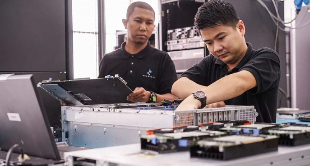 Two male employees dressed in black polo shirts with the Procurri logo reach inside electrical equipment.