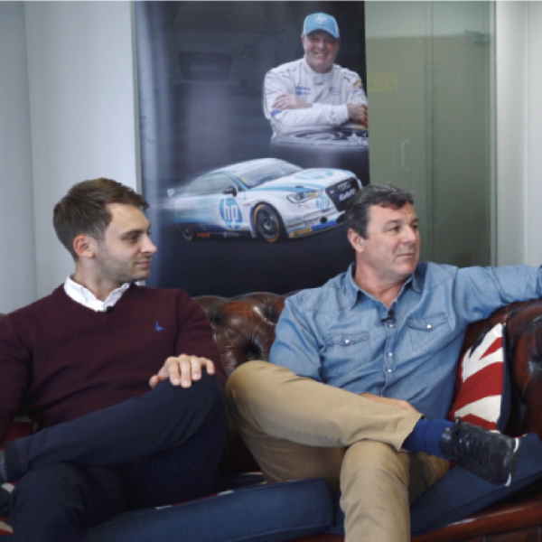 Two men sit cross legged on a sofa below a picture on a wall of a rally driver and car.