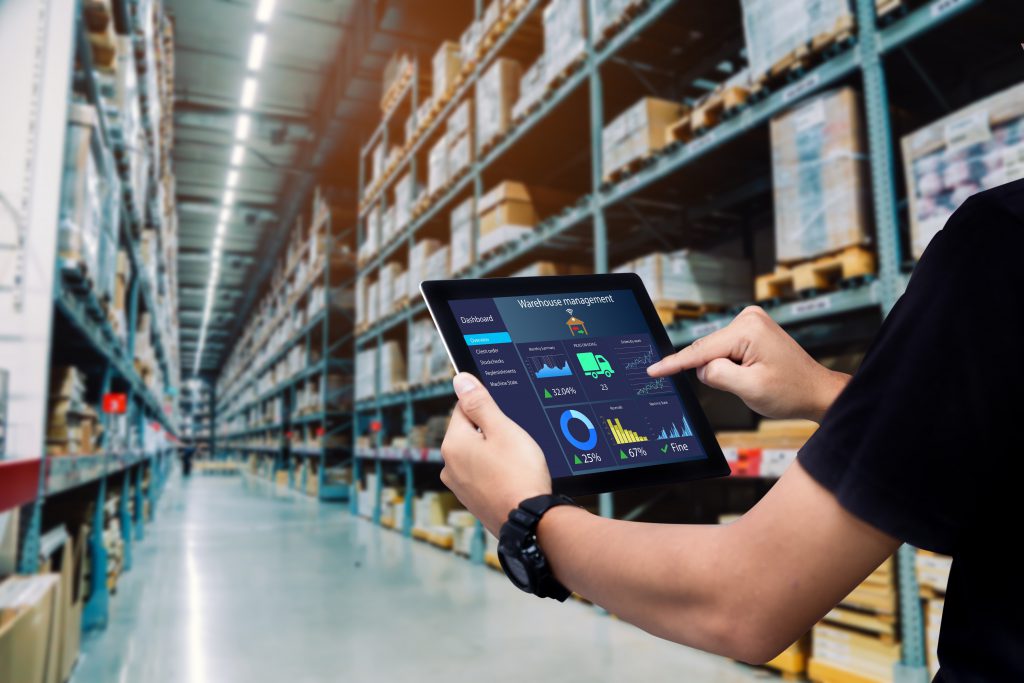 Hands grasp an iPad with a Warehouse Management dashboard on the screen. Cardboard boxes in a warehouse appear behind.