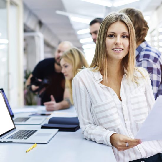 A woman leaning against a desk with a laptop on smiles at the camera as a group of people talk in the background.
