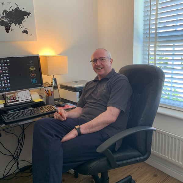 A man sits in an office chair in front of a desktop computer and smiles at the camera.