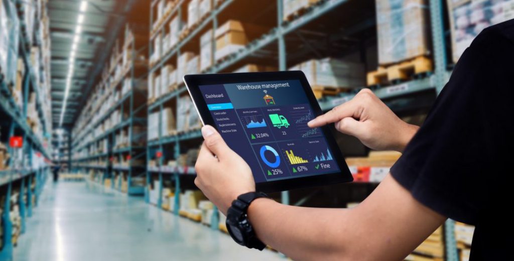 Hands grasp an iPad with a Warehouse Management dashboard on the screen. Cardboard boxes in a warehouse appear behind.