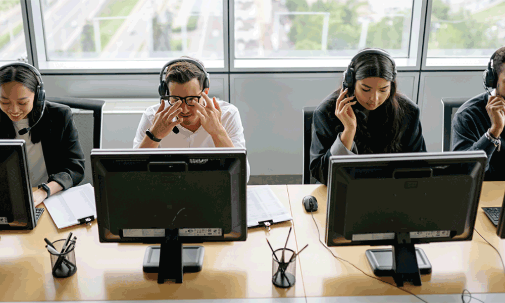 ps-2-2 A row of four people sit in front of screens on a desk wearing headsets. Some are smiling, some look exasperated.