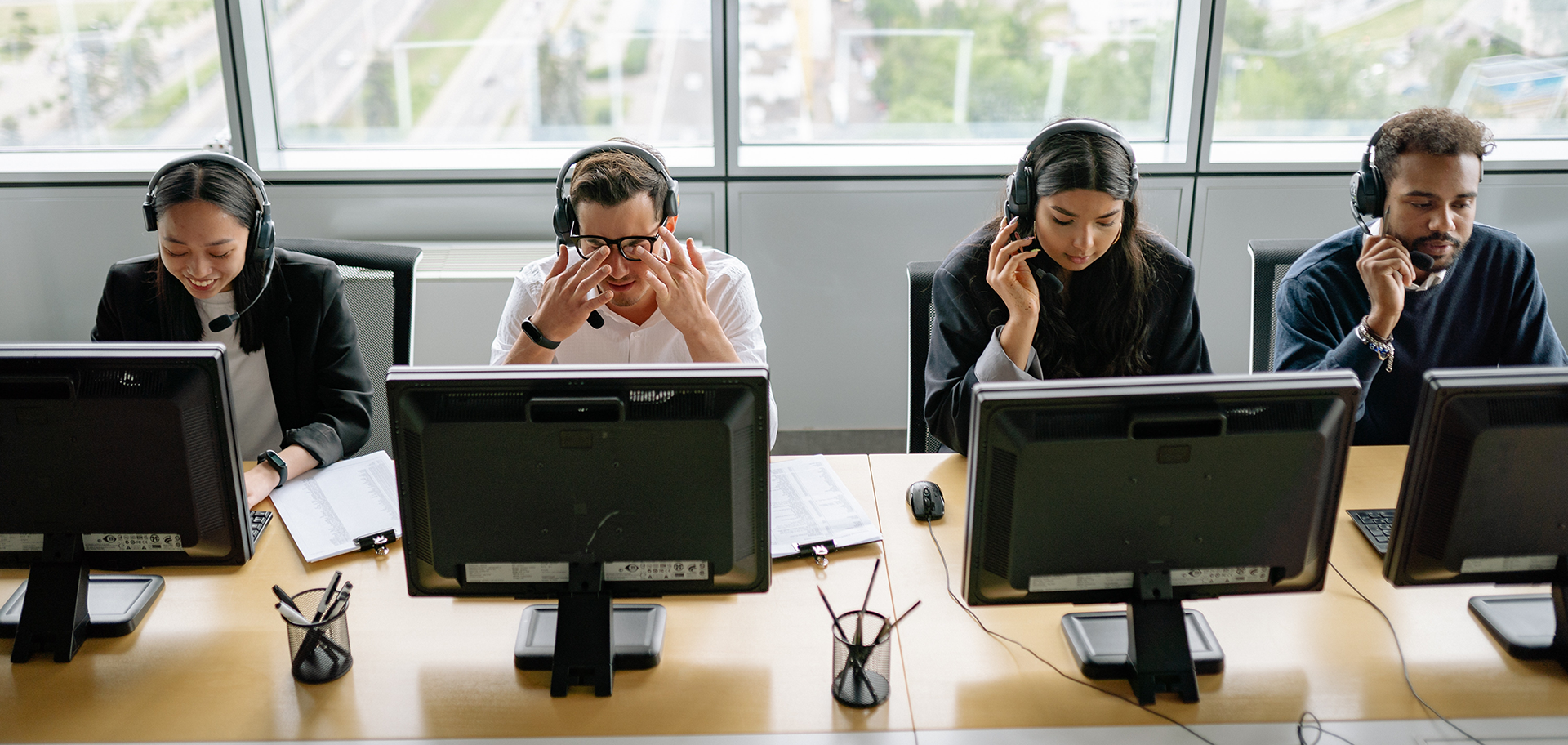 Eine Reihe von vier Personen sitzt mit Kopfhörern vor Bildschirmen auf einem Schreibtisch. Einige lächeln, andere sehen verzweifelt aus.