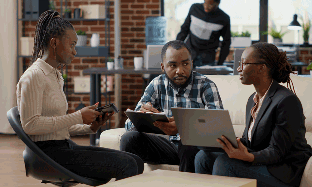 Three adults are sitting on a sofa and stool chatting against a modern office backdrop. Each holds a device; one smartphone, one tablet, one laptop.