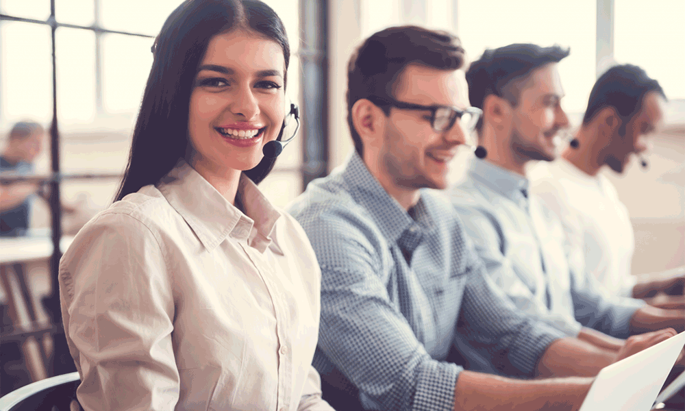 A row of four adults sit next to each other in front of computers, each wearing a headset. The female closest to the camera looks at it and smiles.