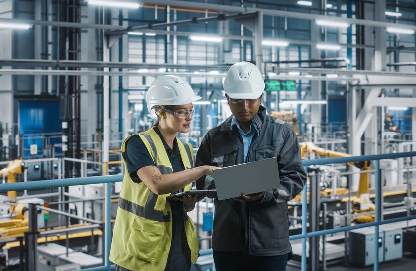 A male and female both dressed in white hard hats against an industrial backdrop look at a laptop and tablet device.