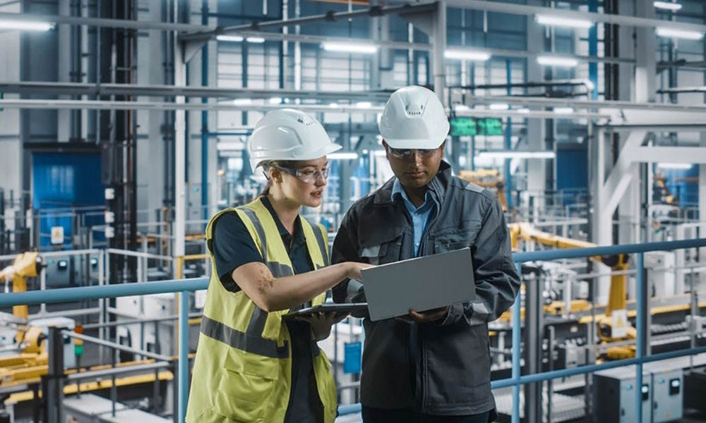 Male,Specialist,And,Female,Car,Factory,Engineer,Talking,And,Working A male and female both dressed in white hard hats against an industrial backdrop look at a laptop and tablet device.