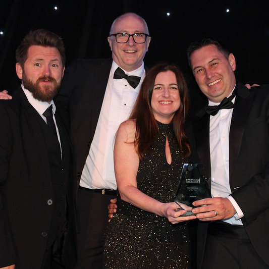 Four Procurri employees, three male and one female, all dressed in black tie face the camera holding a trophy and smiling.
